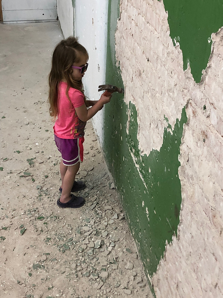 A three year old little girl in pink clothes and purple sunglasses hammers green plaster of the wall to expose the brick beneath